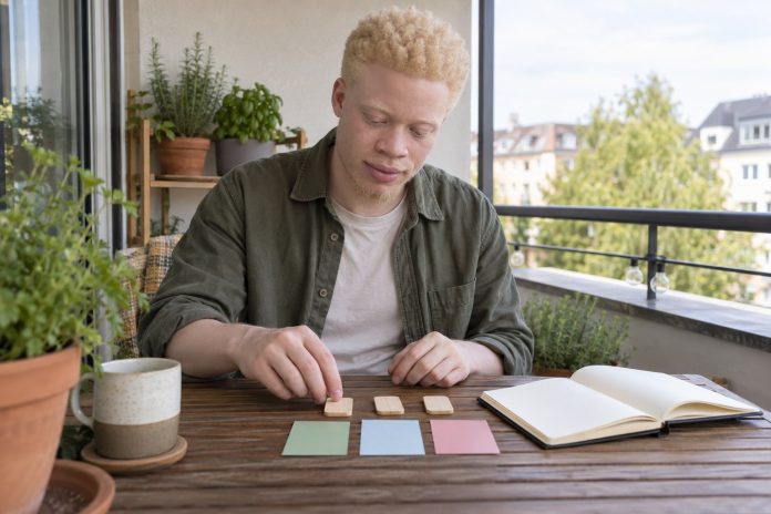 Young Black man with albinism arranging three blank wooden study tiles and colored position cards beside a notebook on a balcony