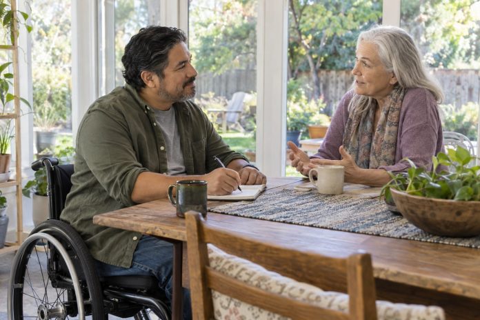Wheelchair user listening and taking notes while an older adult speaks at a sunny home table